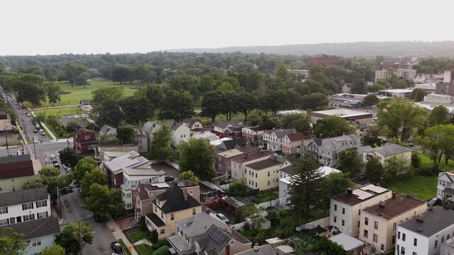 Dense row of gabled colonial homes lines tree shaded street in Orange, New Jersey, parked cars and distant green playing fields stretch beyond hazy town rooftops nearby. Aerial drone rising shot