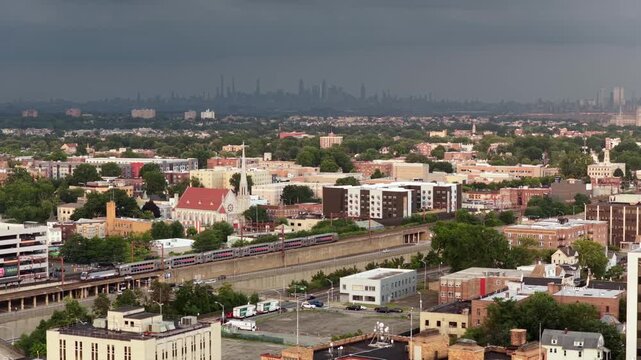 Thunderstorm clouds gather over Orange, New Jersey civic buildings and clock tower, with hazy Manhattan skyline and One World Trade Center looming in the distance. Aerial drone wide shot