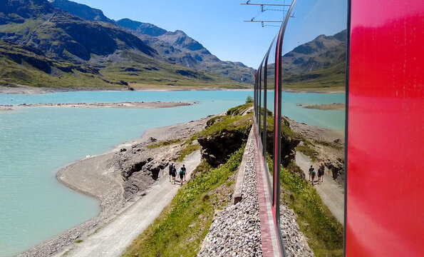 View of the Bernina Express train passing by a turquoise alpine lake with mountains in the background and people walking on a path in Chur, Graubunden, Switzerland.