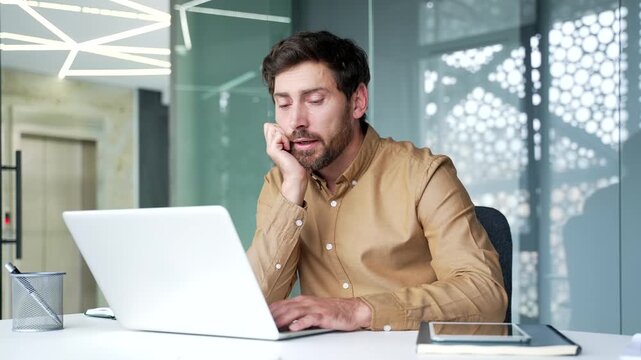 Tired mature businessman looking bored while working on a laptop at his desk in a modern office. Yawning professional struggling with routine tasks during a long business workday at the workplace.
