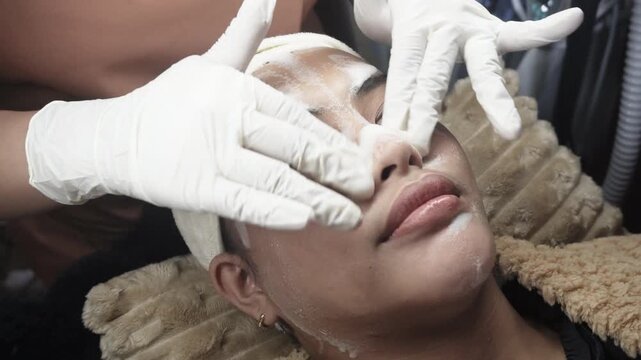 Aesthetician cleansing skin with facial foam during professional deep cleansing treatment at aesthetic clinic for Southeast Asian woman in her early 20s