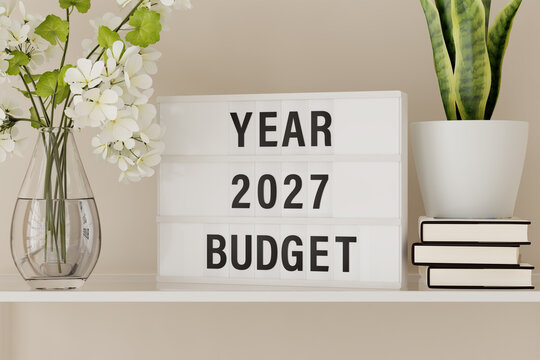 A lightbox displaying the words "Year 2027 Budget" on a shelf with books and plants.