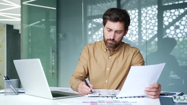 Mature businessman filling out a business form while holding a document in his hands at his desk in a modern office. Focused professional carefully verifying information before completing paperwork.