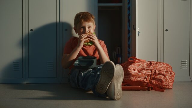 Primary School Boy Sitting on Hallway Floor beside Metal Lockers, Enjoying Burger from Lunch Container During School Break. Childhood, Nutrition, Education, and Everyday Student Life Concept.