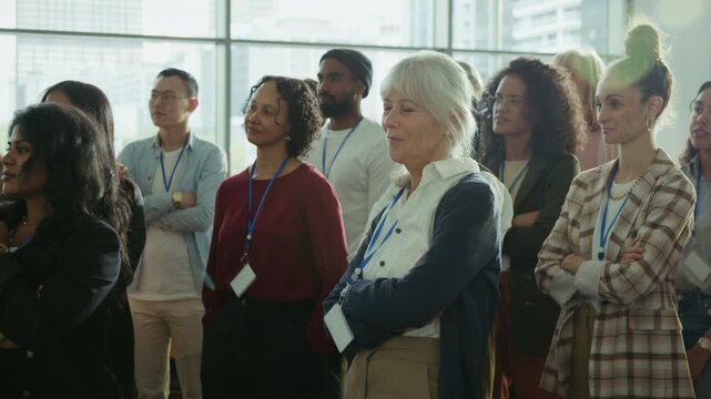 A group of attentive business professionals and individuals stand together, focusing on a speaker at an indoor corporate conference or large event. They are engaged in learning and collaboration.