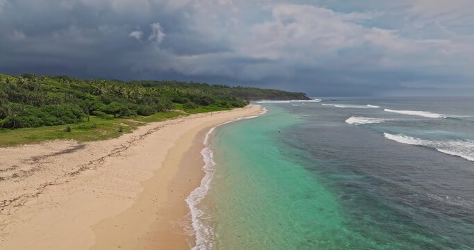 Aerial view of a pristine tropical beach with turquoise waters, gentle waves, and lush green forest under a dramatic cloudy sky in Port Resolution, Tanna Island, Vanuatu.
