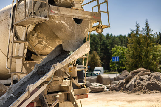 A mixer truck is pouring concrete at a construction site near a road. Construction materials and equipment are scattered around the area.