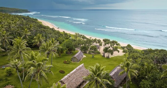 Aerial view of traditional thatched-roof huts nestled among lush palm trees along a pristine tropical beach with turquoise water and rolling waves in Port Resolution, Tanna Island, Vanuatu.