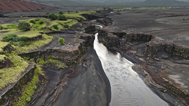 Aerial view of a winding river flowing through a volcanic landscape with black sand, rugged rock formations, and patches of vibrant green vegetation in Port Resolution, Tanna Island, Vanuatu.