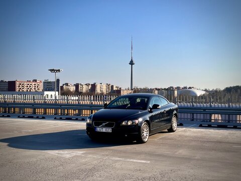 A black second-generation Volvo C70 convertible is parked on a rooftop with a modern cityscape in the background. The setting is Vilnius, Lithuania, recognizable by the Vilnius TV Tower visible 