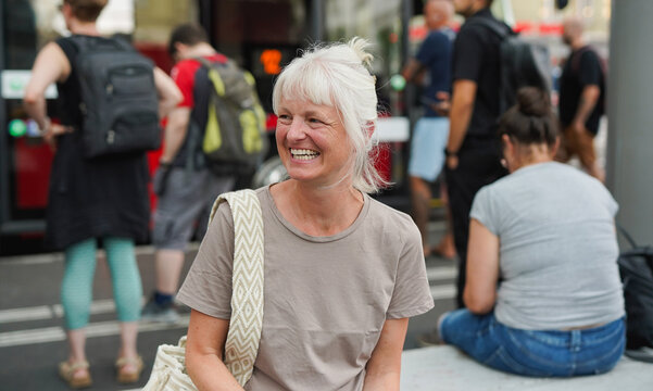 Happy senior woman laughing while waiting at bus station in the city - Travel, lifestyle and mature person concept