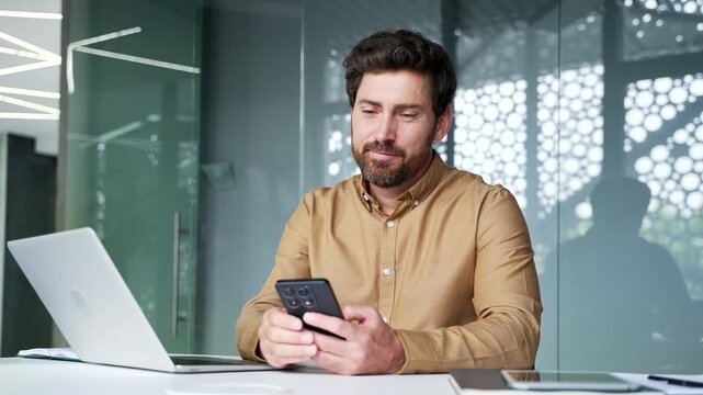 Smiling mature businessman browsing a smartphone while sitting at his workplace in a modern office. Happy professional checking mobile content at his desk during a productive business day.