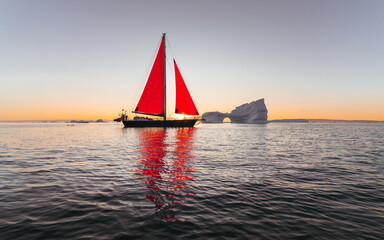 Sailboat with striking red sails cruising among icebergs near Ilulissat, Greenland, during the golden light of the midnight sun. Scenic Arctic seascape with floating ice, calm water and colorful refle © Dagmar