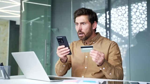 Shocked mature businessman holding a credit card and smartphone while sitting at workplace in a modern office. Worried professional discovering unauthorized banking charges and suspected payment fraud