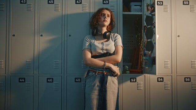 Teenage Schoolgirl With Headphones at Neck Standing in High School Locker Room. Girl Holds Smartphone, Staring at Screen With Upset Facial Expression While Reading Text Message or Social Media Post.