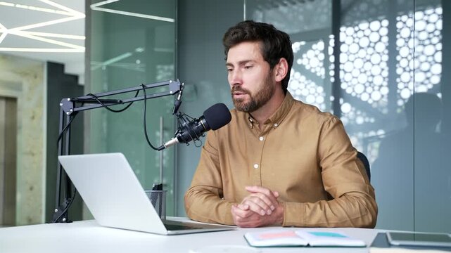 Mature businessman wearing headphones speaking into a microphone while using a laptop in a modern office. Professional host recording a business podcast, webinar, or online training session.