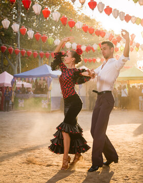 A young couple performs a synchronized sevillanas dance step on the dusty fairground under rows of colorful paper lanterns.