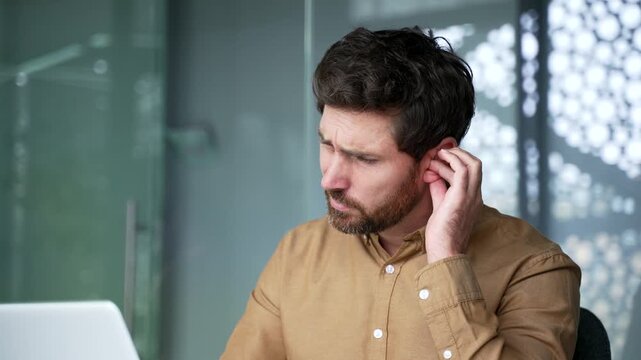 Close-up of a mature businessman touching his painful ear at workplace in a modern office. Sick professional experiencing severe earache caused by an ear infection or inflammation during the workday.