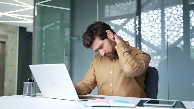 Mature businessman rubbing his painful neck while working on a laptop at his desk in a modern office. Tired professional experiencing muscle stiffness and discomfort after long hours of computer work.