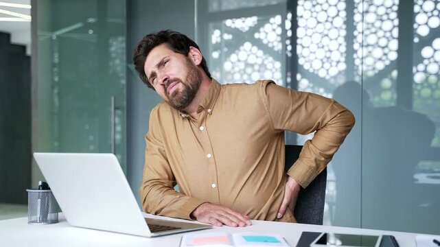 Mature businessman feeling lower back pain while sitting at his desk at workplace in a modern office. Uncomfortable professional rubbing his sore back to relieve muscle tension during the workday.