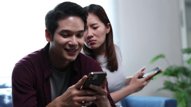Jealous girlfriend secretly looks at boyfriend's smartphone screen while they sit together on the living room sofa.
