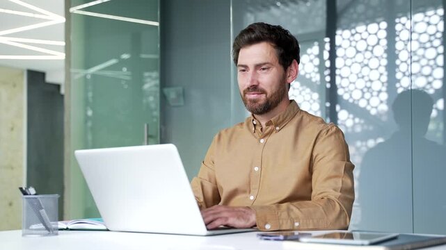 Smiling mature businessman using a laptop while working at his desk in a modern business office. Confident professional focused on daily tasks in a corporate workplace with a positive attitude.