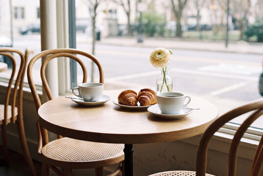 Romantic cafe table for two with white coffee cups croissants and single flower in soft morning light