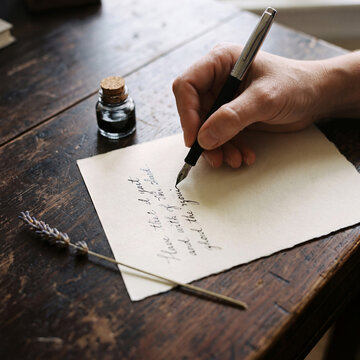 Overhead view of hand writing cursive letter with fountain pen on cream paper with inkwell and dried flower
