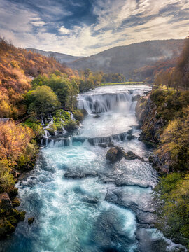 Aerial view of Strbacki buk Waterfall with vibrant autumn forests and rocky riverbanks under a cloudy sky in Orasac, Federation of Bosnia and Herzegovina, Bosnia and Herzegovina.