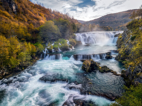 Aerial view of Strbacki buk waterfall cascading over rocks into the turquoise Una River, surrounded by autumn forests in Orasac, Federacija Bosne i He, Bosnia and Herzegovina.