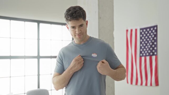 Man young points finger to i voted sticker on chest with american flag hanging on wall in studio; pride civic duty.