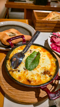 Vertical close-up shot of hot traditional baked cheese dish with golden melted crust garnished with fresh green leaf, served in a red cast iron skillet on a wooden board.