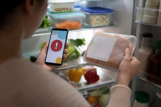 Woman holding packaged raw chicken and checking a food recall alert on her smartphone in front of an open refrigerator.