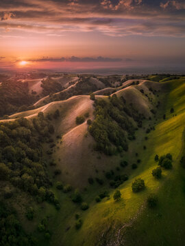 Aerial view of the rolling green hills of Zagajicka brda at sunset with golden light illuminating the grassy slopes and scattered trees in Vrsac, Vojvodina, Serbia.