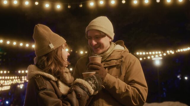 Caucasian young couple standing outside at night under hanging string lights, drinking hot beverages from paper cups and laughing. Footage is suitable for winter holiday marketing campaigns