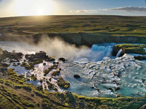 Aerial view of the majestic Godafoss Waterfall at sunset, with mist rising from the cascading water over rocky terrain amidst the lush green landscape of Godafoss, Thingeyjarsveit, Iceland.