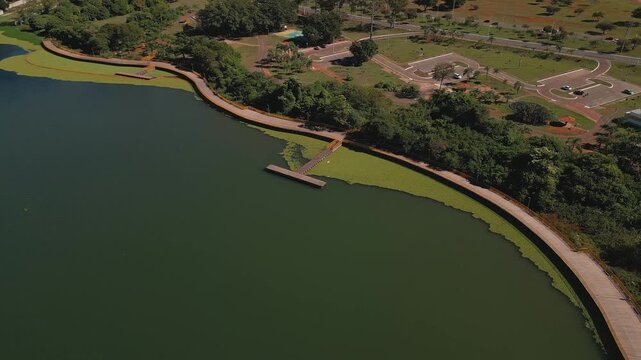Drone performs a smooth parallax orbit above Lake Paranoa, Brasilia, Federal District, Brazil, revealing extensive floating invasive vegetation, lakeside park, wooden pier and the urban waterfront.
