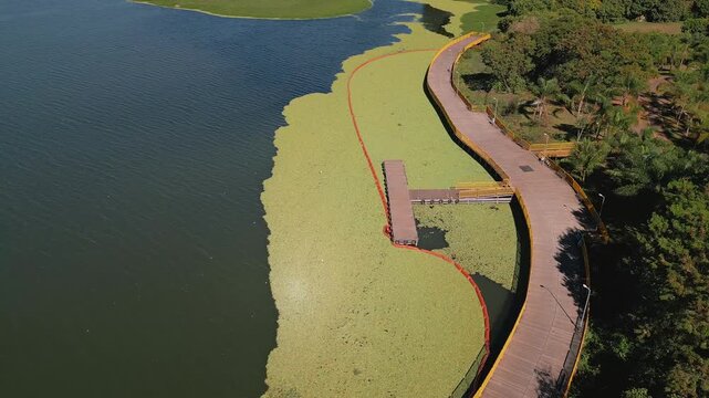 Drone performs a parallax orbit above Lake Paranoa, Brasilia, Federal District, Brazil, revealing a wooden pier, floating water hyacinth, containment boom, lakeside promenade and open freshwater.