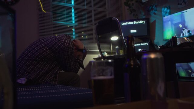 Young adult Caucasian man sitting in dark room holding head in hands near gaming screens, experiencing stress and screen addiction. Footage suitable for mental health and digital burnout campaigns