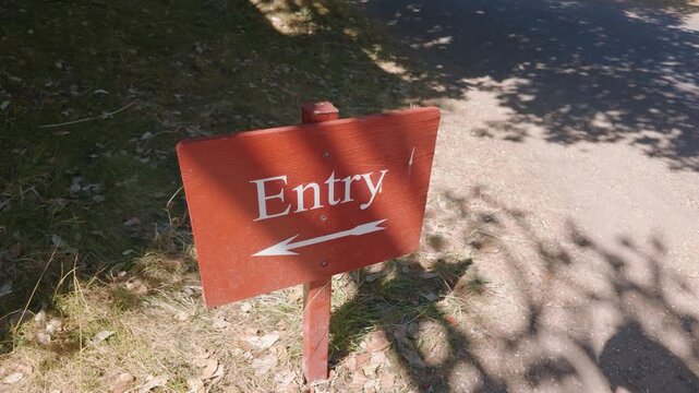 Entry sign with left arrow on orange wooden board as camera pans with slight orbit. Dappled shade, gravel path and grass suggest park or venue entrance wayfinding.