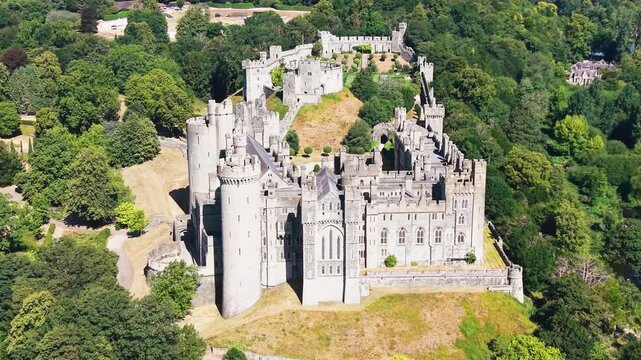 Arundel castle aerial orbit reversing as drone backs away from medieval fortress and towers, revealing wooded hillside parkland in West Sussex, England on a bright summer day.