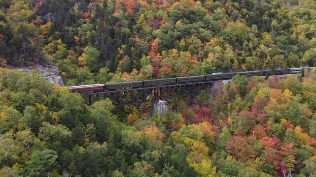 Drone aerial view of a vintage passenger train crossing the historic Willey Brook Trestle bridge in Crawford Notch, New Hampshire. Surrounded by vibrant peak autumn foliage and mountain forest scenery