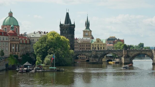 Historic Charles Bridge spans the Vltava River in Prague near the Old Town bridge tower. Small tour boats navigate the water under sunny skies. Classic European architecture highlights tourism.