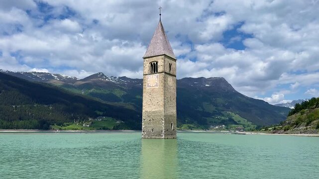 Locked-off view of the submerged Curon bell tower in turquoise Lake Resia, beneath moving clouds in early summer.
