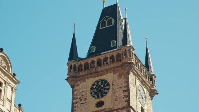 Tourists stand on the observation deck of the Old Town Hall tower in Prague. Historic stone clock tower features gothic architecture under a clear sky. Iconic landmark for European travel.
