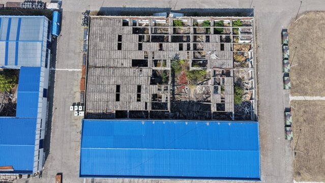 Aerial view of the Severna Zona industrial area in Pancevo, featuring a derelict building with a partially collapsed roof and vegetation next to a modern blue-roofed facility in Pancevo, Serbia.