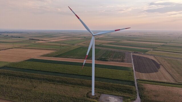 Aerial view of a wind turbine standing in a vast agricultural field with patchwork crops under a soft sunset sky in Crepaja, Vojvodina, Serbia.