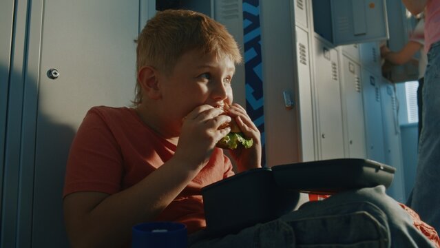 Young Schoolboy Sitting Near Metal Lockers Having Lunch in Bright School Corridor With Reusable Food Container and Insulated Bottle Nearby. Authentic Education, Nutrition, and Daily Student Routine.