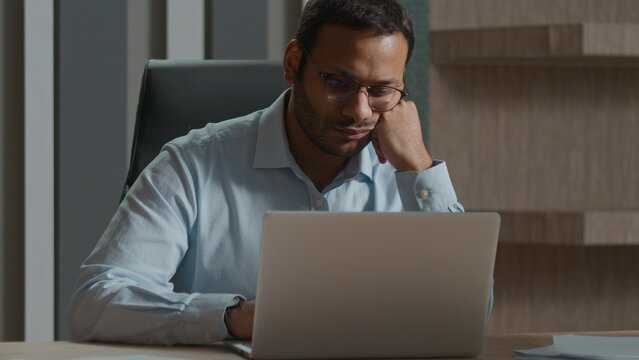 Tired Indian Arabian business man yawning in office bored with computer laptop work evening deadline exhausted businessman manager fatigued no energy overworked napping after online job stress burnout