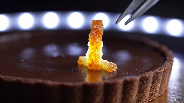 Hand using tweezers to place vibrant orange candy decoration on a chocolate tart, showcasing meticulous attention to detail in a well-lit kitchen setting